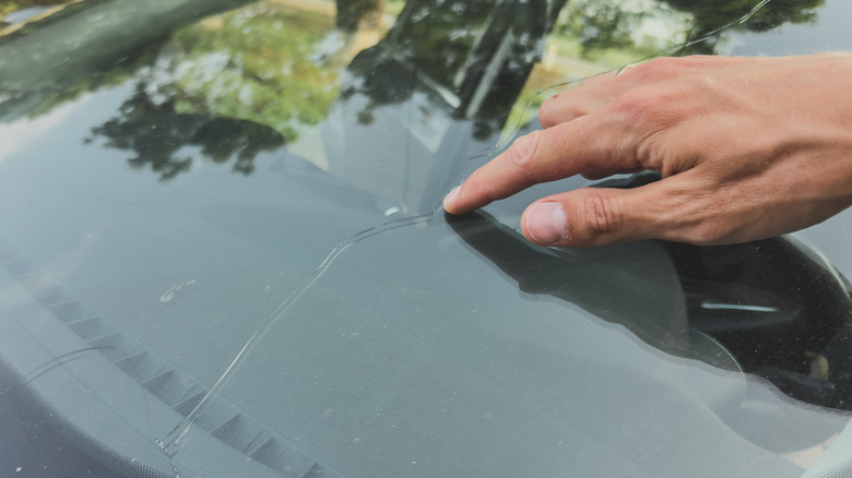 A person's hand pointing at a long crack in a car windshield, highlighting glass damage.