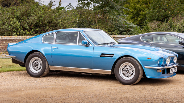 A light blue 1977 Aston Martin Vantage parked in a country setting