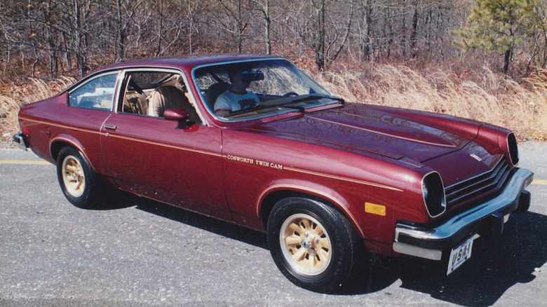 A maroon 1976 Chevrolet Cosworth Vega twin-cam parked on asphalt in front of bare-limbed trees
