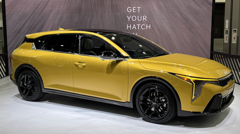 A front three-quarters shot of a gold kia K4 Hatchback on a white rotating platform in front of a screen that reads, 