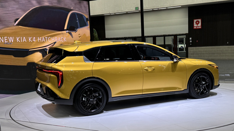 Rear three quarters shot of a gold Kia K4 Hatchback parked on a white rotating platform at the LA Auto Show in front of a screen that says "The All-New Kia K4 Hatchback"