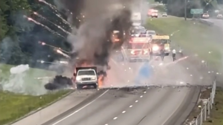 Fireworks explode from a burning trailer on the side of the interstate as firefighters wait and block traffic from a safe distance