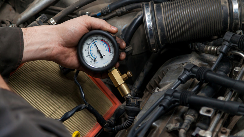 A mechanic reading the results of a compression test on a vehicle engine, with a pressure gauge indicating roughly 150 PSI.