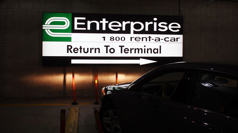 A backlit Enterprise rental car sign  in a parking garage directs cars (like the sedan pictured) to make a right to return to the terminal.