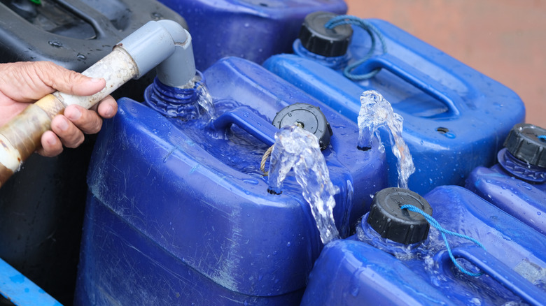 A hand fills multiple blue water jugs from a hose.
