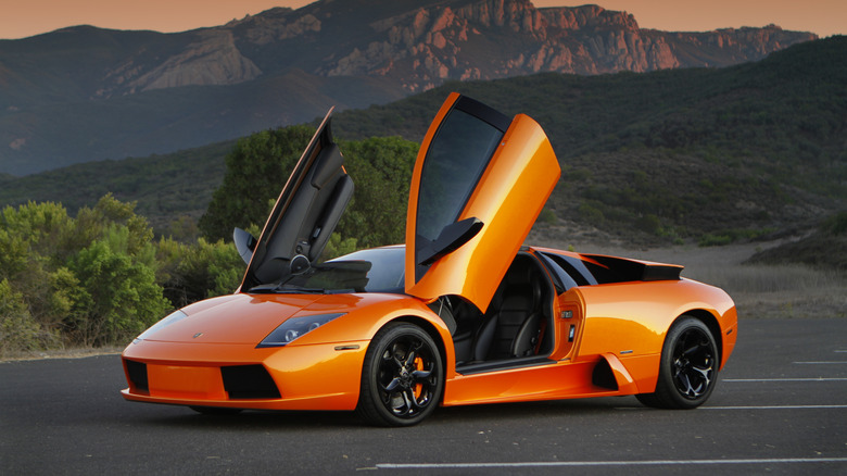 An orange Lamborghini Murcielago with its doors open in a parking lot in front of green rolling hills and mountains