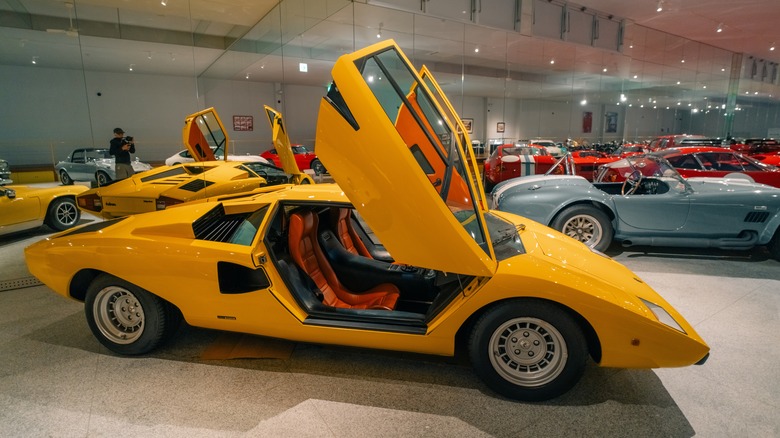 A yellow Lamborghini Countach LP400 sits in a museum collection with its doors open, while a second LP400 and a Shelby Cobra sit behind it