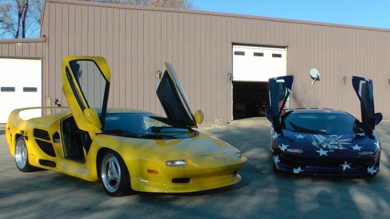 A yellow Vector M12 next to an America-themed wrapped Vector M12, both parked with their doors open in front of a brown building