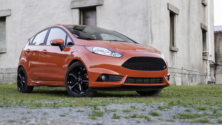 A reddish-orange Ford Fiesta ST four-door hatchback parked in front of an abandoned building