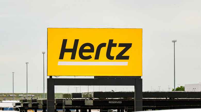 A Hertz rental parking lot sign with street lights in the background.