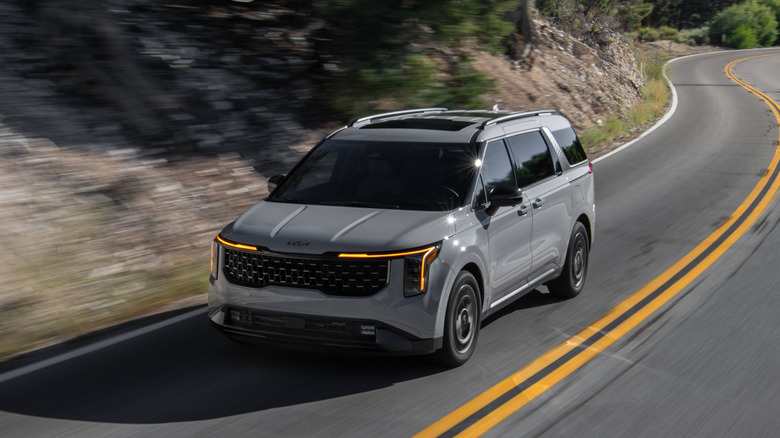 A high-angle action shot of a 2026 Kia Carnival Hybrid in Ceramic Silver navigating a winding mountain road.