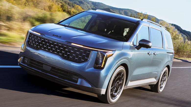 A low-angle, front-three-quarter view of a 2026 Kia Carnival in Astra Blue driving on a coastal highway at sunset.