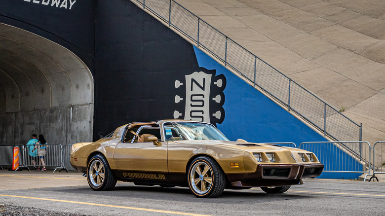 1979 Pontiac Firebird Formula gold convertible driving out of a tunnel at a racetrack.
