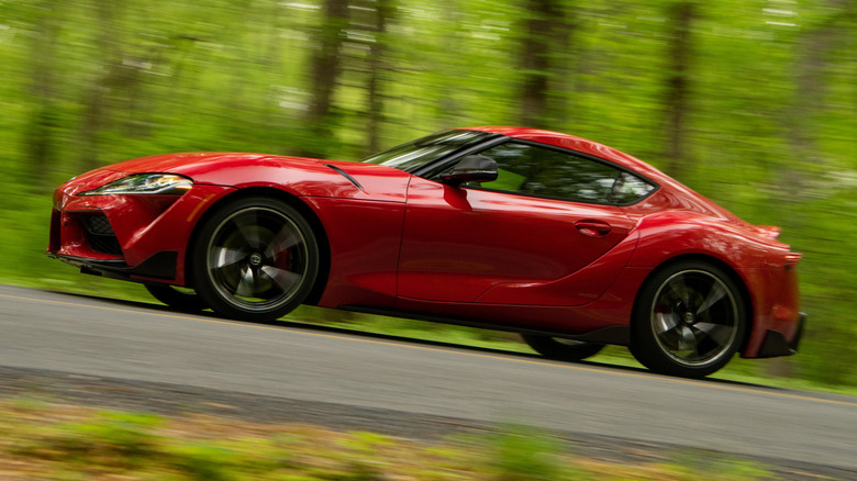A bright red Toyota Supra A90 in motion on road past a wooded area.