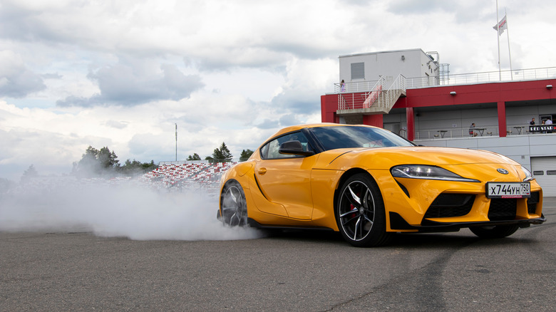 A yellow Toyota Supra with tire smoke coming out the back and racetrack grandstand in the background.