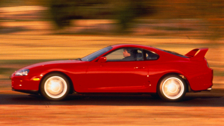 A red Toyota Supra A80 viewed in profile at speed, with a driver at the wheel.