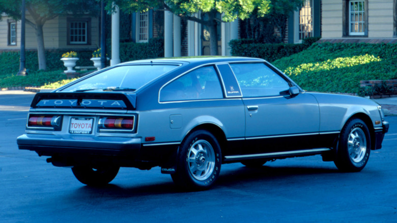 1981 Toyota Celica Supra viewed from rear three quarters in grey and back parked in leafy street with buildings in the background.