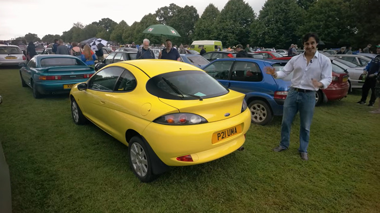 screenshot from a YouTube video showing a rear three quarters shot of a bright yellow Ford Puma parked on grass with a man standing next to it in front of other classic cars