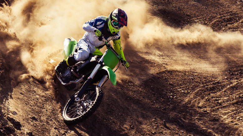A rider makes a turn riding a green dirtbike through a rock-covered landscape with dust billowing up behind