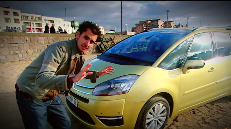 a screenshot of Jonny Smith standing in front of a yellow Citroen C4 Picasso parked on the beach