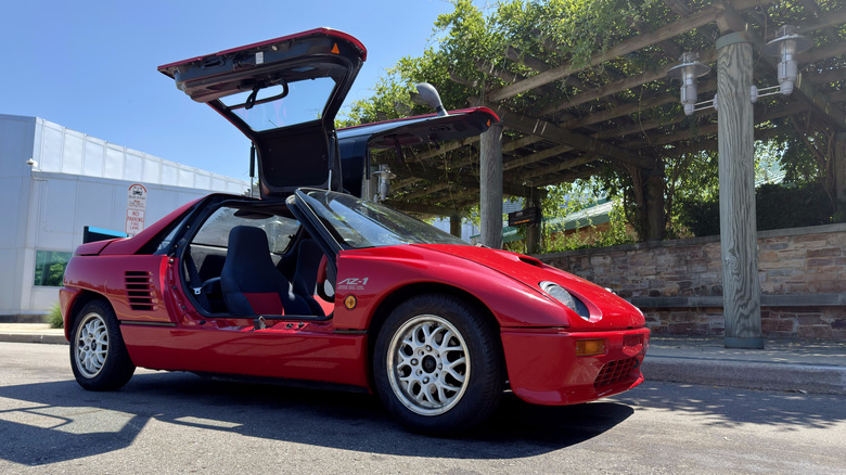 A red Mazda Autozam AZ-1 with its gullwing doors opened, parked at the front of a building with a green-laden trellis out front.