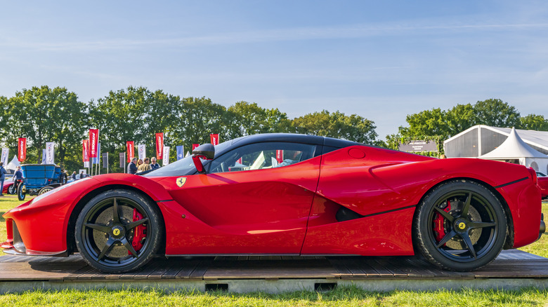 A side view of a black and red Ferrari LaFerrari parked in the sun with greenery and other cars on display.