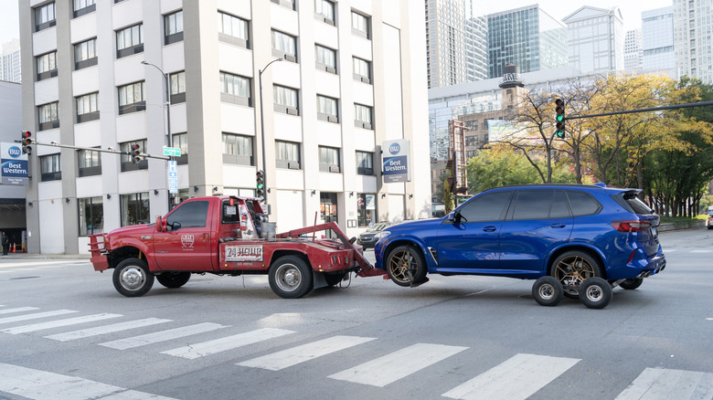 A blue BMW SUV is pulled through a city by a red wheel-lift tow truck.