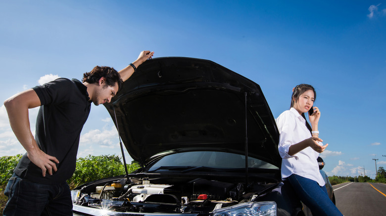 A man looks at the engine bay of a broken down car while a woman is on the phone calling for assistance.