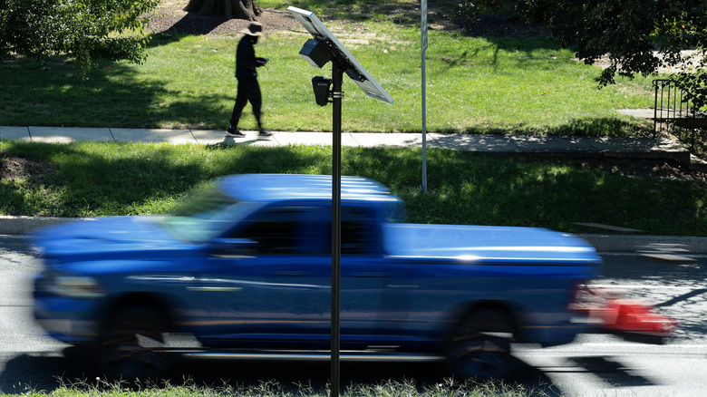 An Axon ALPR camera and solar panel mounted on a pole next to a road as a blue pickup truck and pedestrian on a sidewalk go by