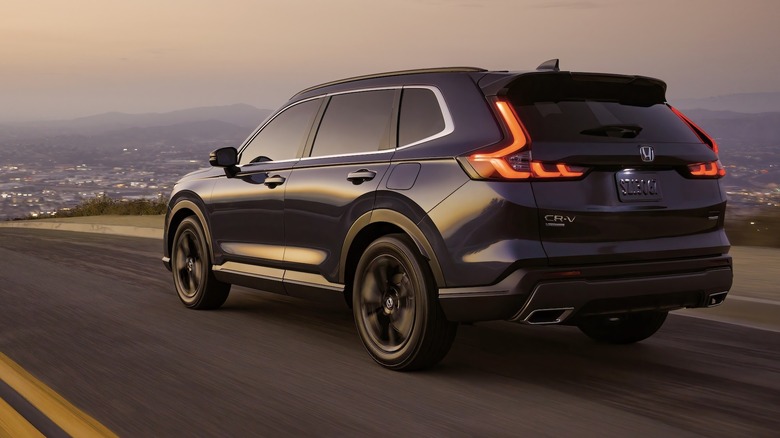 Rear 3/4 view of a black 2024 Honda CR-V driving down a road at dusk with a town and mountains in the background