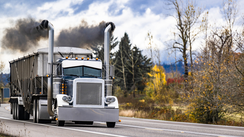 Big rig black classic semi truck with extended cab for truck driver rest and black smoke from the exhaust pipes polluting the environment transporting cargo in bulk semi trailer running on the road