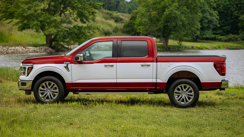 Side view of a red 2027 F-150 with a white stripe down the side parked on grass in front of a lake and trees