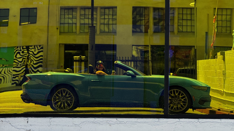 A side view of me in the driver's seat of the bright blue Mustang EcoBoost with the top down taken in the reflection of a shiny yellow-tinted window