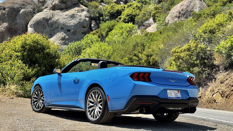 A rear three-quarters shot of the bright blue Mustang EcoBoost with its top down  parked on gravel in front of a shrub-covered mountainside