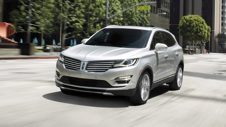 Front three quarters shot of a silver 2016 Lincoln MKC driving through a city in front of trees and buildings