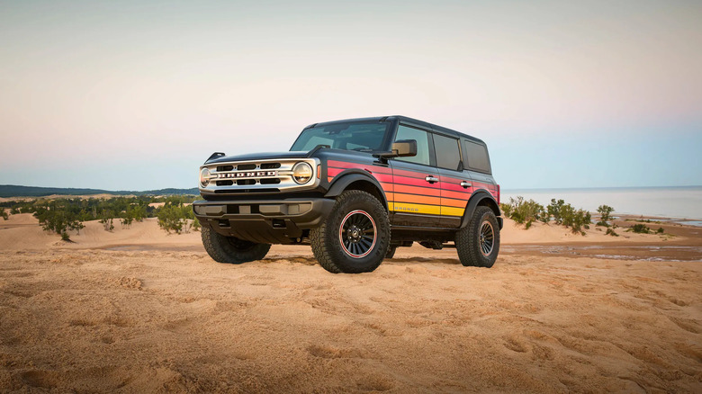 A Ford Bronco sits dramatically on the beach with a rainbow color scheme and water behind.