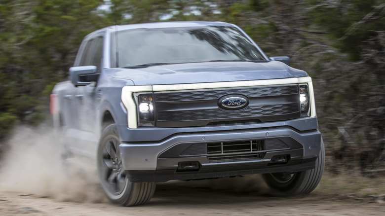 A head-on shot of a light blue F-150 Lightning driving on a dirt road and kicking up dust in front of trees