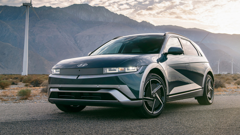 A front three-quarters shot of a blue Ioniq 5 parked on asphalt in front of wind turbines in the desert and the silhouette of a mountain range at sunset