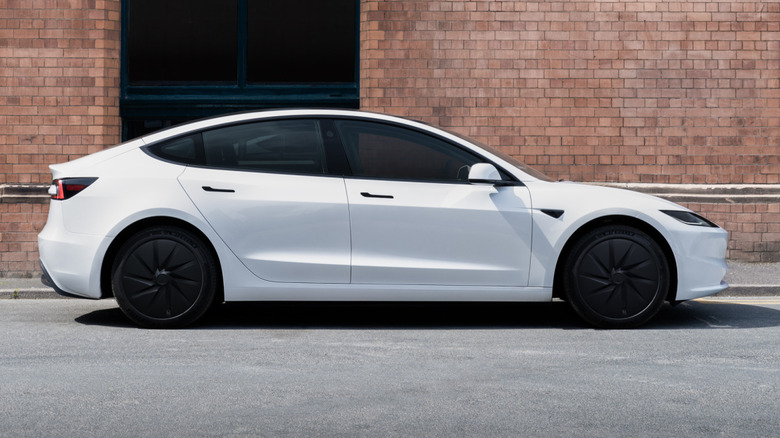 A side view of a white Tesla Model 3 Rear-Wheel-Drive with black wheels and black door handles in front of a brick building
