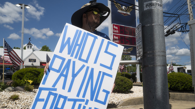 A man in a floppy large sun hat stands in front of a Freedom Fuel gas station, with a sign that reads "Who is paying for this?"