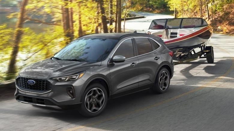 A metallic grey Ford Escape SUV tows a red-and-white motorboat on a trailer down a winding, sun-dappled asphalt road.