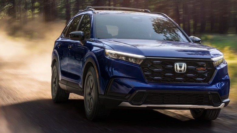 A deep blue 2026 Honda CR-V TrailSport Hybrid drives down an unpaved dirt path, kicking up thick clouds of dust behind it.
