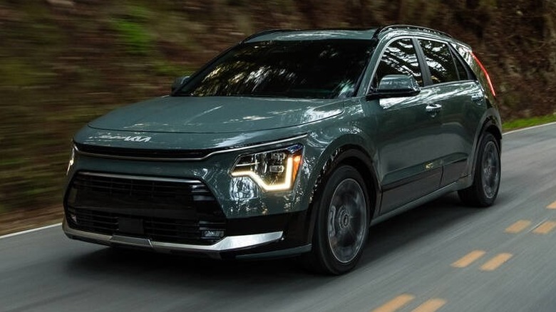 A deep sage green 2026 Kia Niro crossover sweeping through a curve on a two-lane forest road.