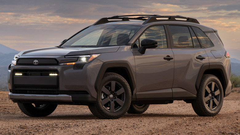 An Urban Rock grey 2026 Toyota RAV4 Hybrid Woodland Edition compact SUV equipped with black off-road wheels, all-terrain tires, and a roof rack.