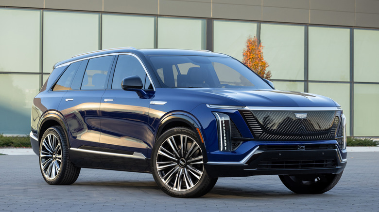 Front three quarters shot of a dark blue Cadillac Vistiq parked in front of a glass building
