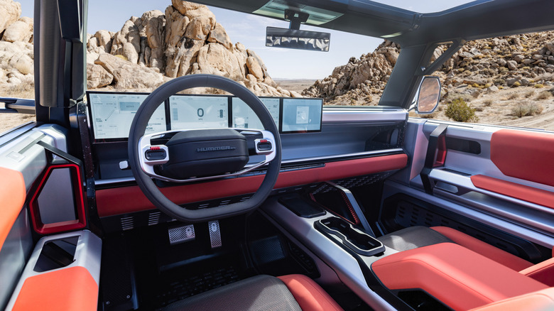 A wide shot of the dashboard of the Hummer X SUV with a red interior, four individual screens on the dashboard, and a rocky desert landscape visible out the windows