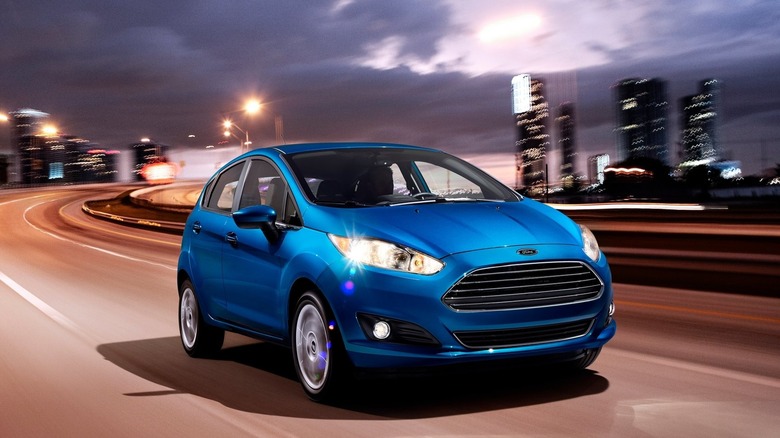 Front three-quarters shot of a blue Ford Fiesta driving away from an urban skyline at dusk