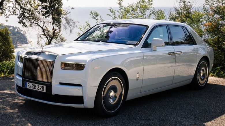 A white Rolls-Royce Phantom parked on a gravel driveway with trees in the background and the ocean behind them