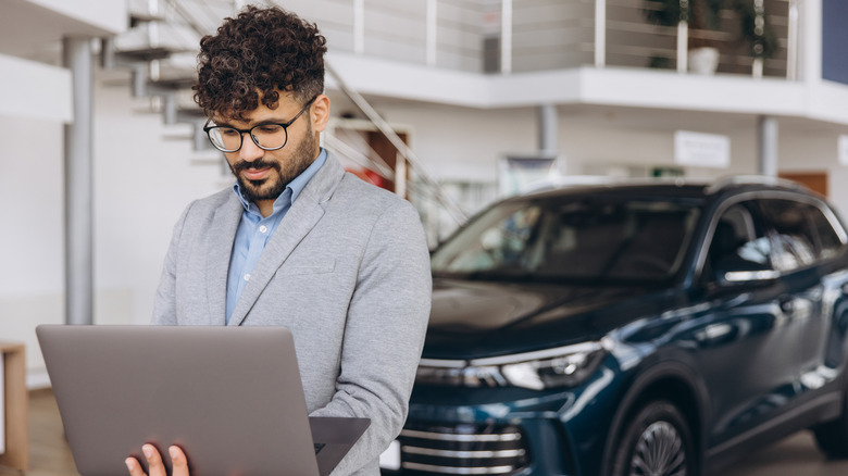 Salesman using laptop in car dealership showroom, consulting data for selling new car to customer