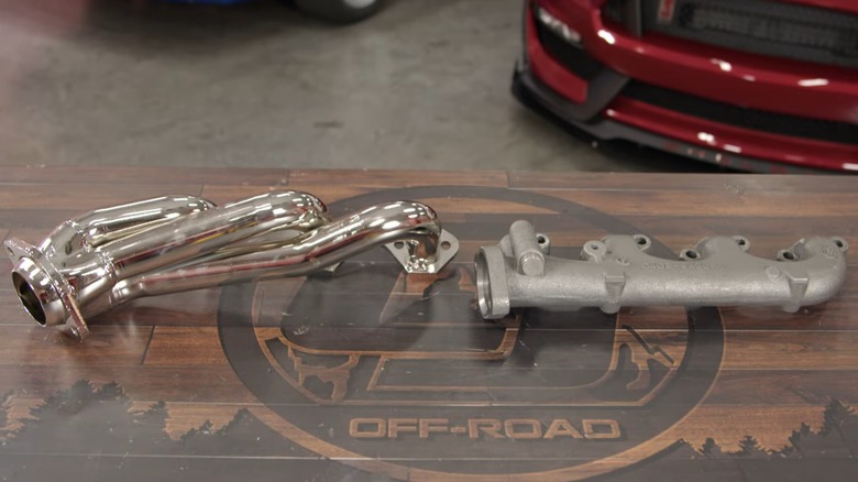 An set of aftermarket exhaust headers next to a stock exhaust manifold on a wooden work bench with a red car in the background